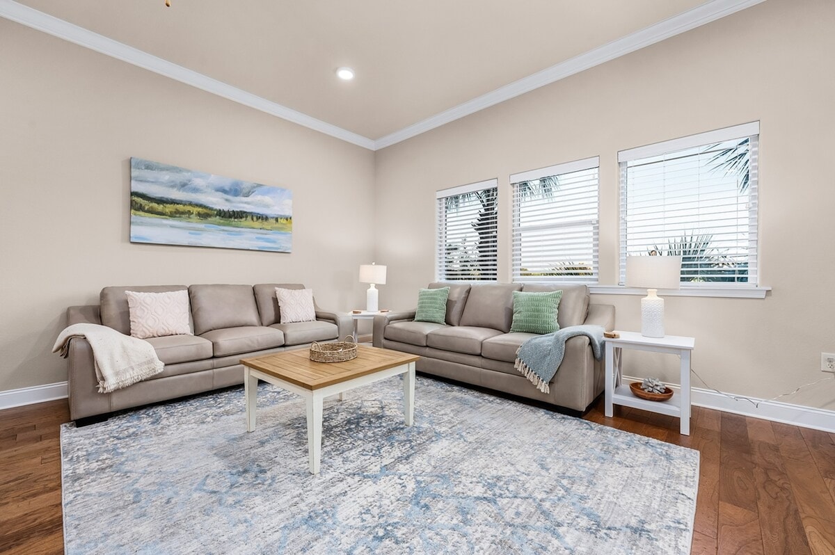 A comfortable living area features two light-colored sofas and a wooden coffee table at the center. Natural light pours in through three large windows adorned with white blinds, illuminating the soft blue and beige tones of the decor. A textured area rug complements the space.