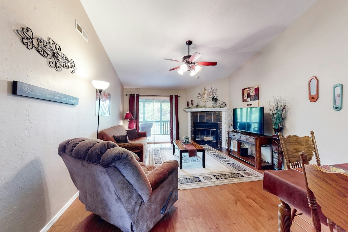 A spacious living area features two comfortable armchairs and a couch, offering a relaxed seating arrangement. A coffee table is centered in front of a television mounted on the wall, with light streaming from the nearby sliding glass doors that lead to a balcony.