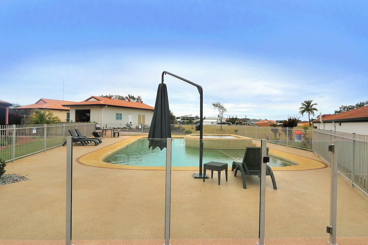 The outdoor area features a fenced-in pool with a gentle curve, surrounded by a spacious deck. Poolside lounge chairs provide seating, while a shaded umbrella stands nearby. Lush greenery and distant structures create a tranquil backdrop.