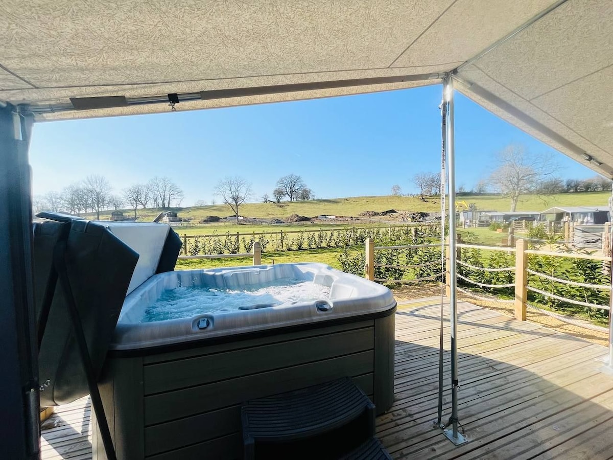 A hot tub is situated on a wooden deck, surrounded by a serene landscape of rolling hills and trees. Clear blue skies create a bright and inviting atmosphere, enhancing the outdoor relaxation experience.