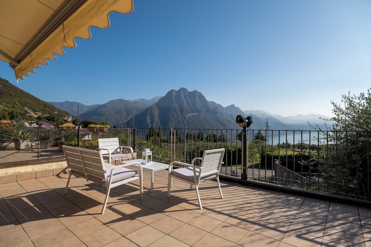 An expansive terrace is featured, offering stunning views of surrounding mountains and Lake Iseo. Comfortable white lounge chairs are arranged around a small table, with an awning providing shade. The space emphasizes outdoor relaxation and scenic beauty.