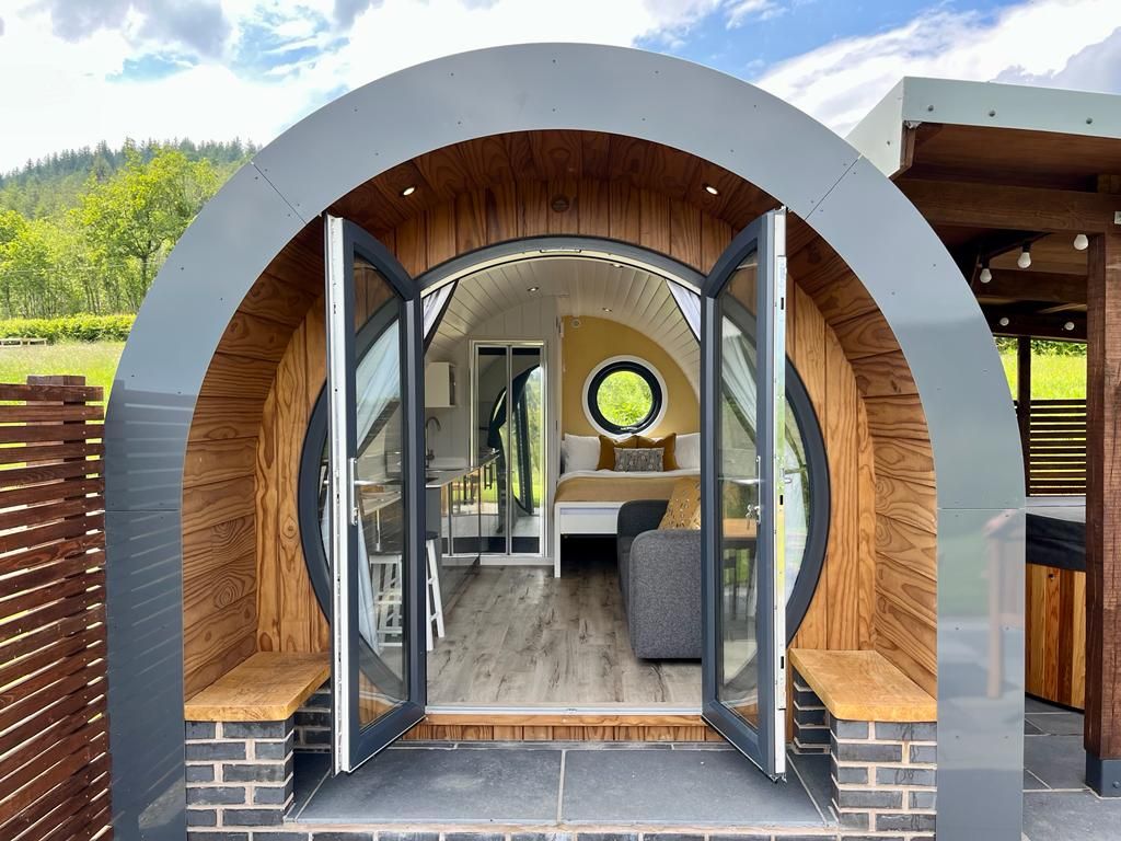 The glamping pod is framed by a curved wooden structure with large open doors, showcasing a bright interior that includes a spacious bed and a cozy seating area. Natural light floods the space, highlighting the modern design and surrounding greenery.