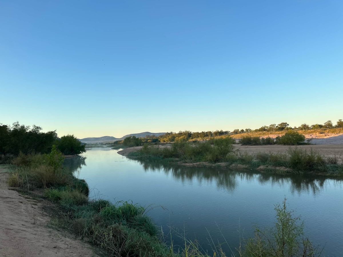 The serene Llano River winds gently through the landscape, reflecting the clear blue sky. Lush greenery frames the riverbanks, while distant hills create a tranquil backdrop, inviting exploration and relaxation in the natural setting.