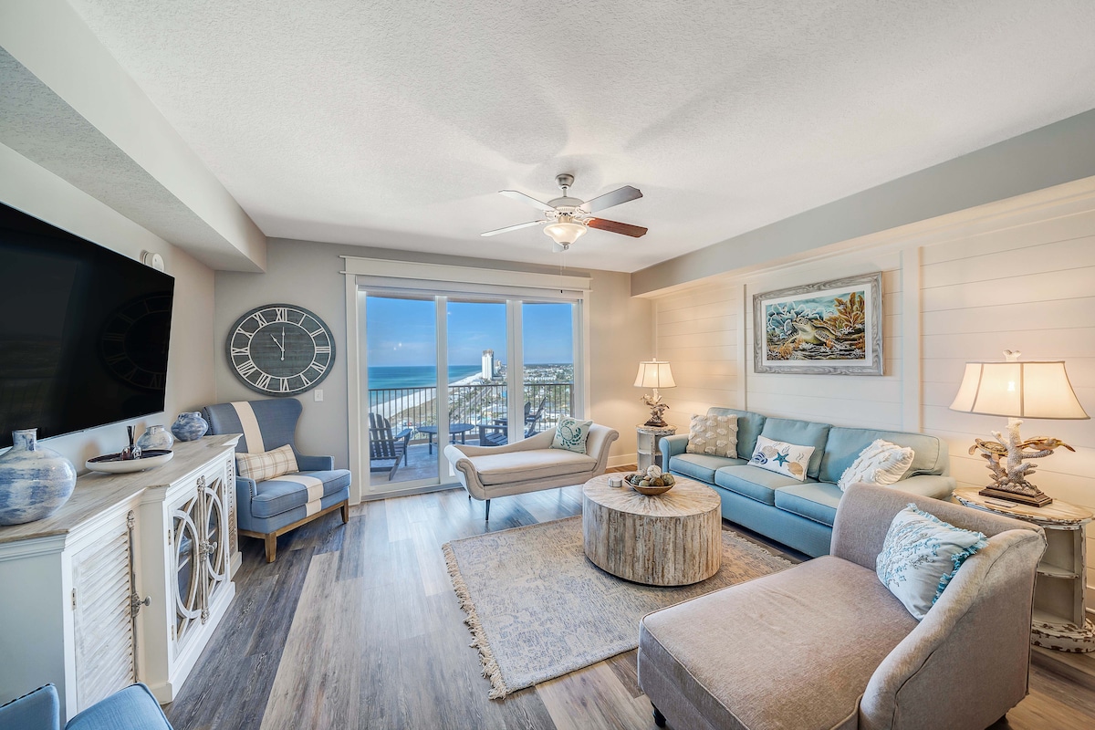 A bright and spacious living area is highlighted by large glass doors leading to a private balcony with ocean views. Comfortable seating arrangements include a stylish sofa and armchairs. A decorative table centers the room, accented by coastal decor and soft blue tones throughout.