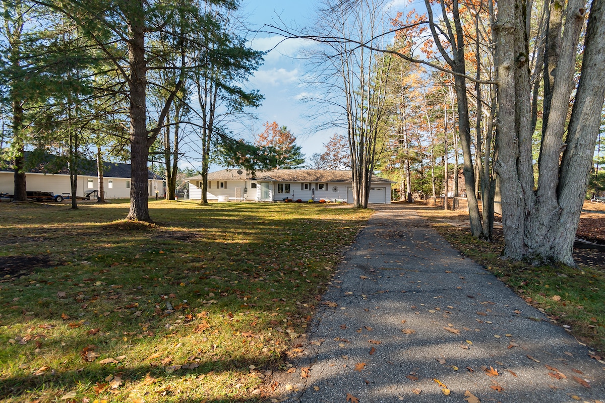 A gravel driveway leads through a wooded area to a single-story home, framed by trees with autumn foliage. The front yard is spacious, featuring a well-maintained lawn with scattered fallen leaves. The backdrop includes neighboring homes set back from the road.