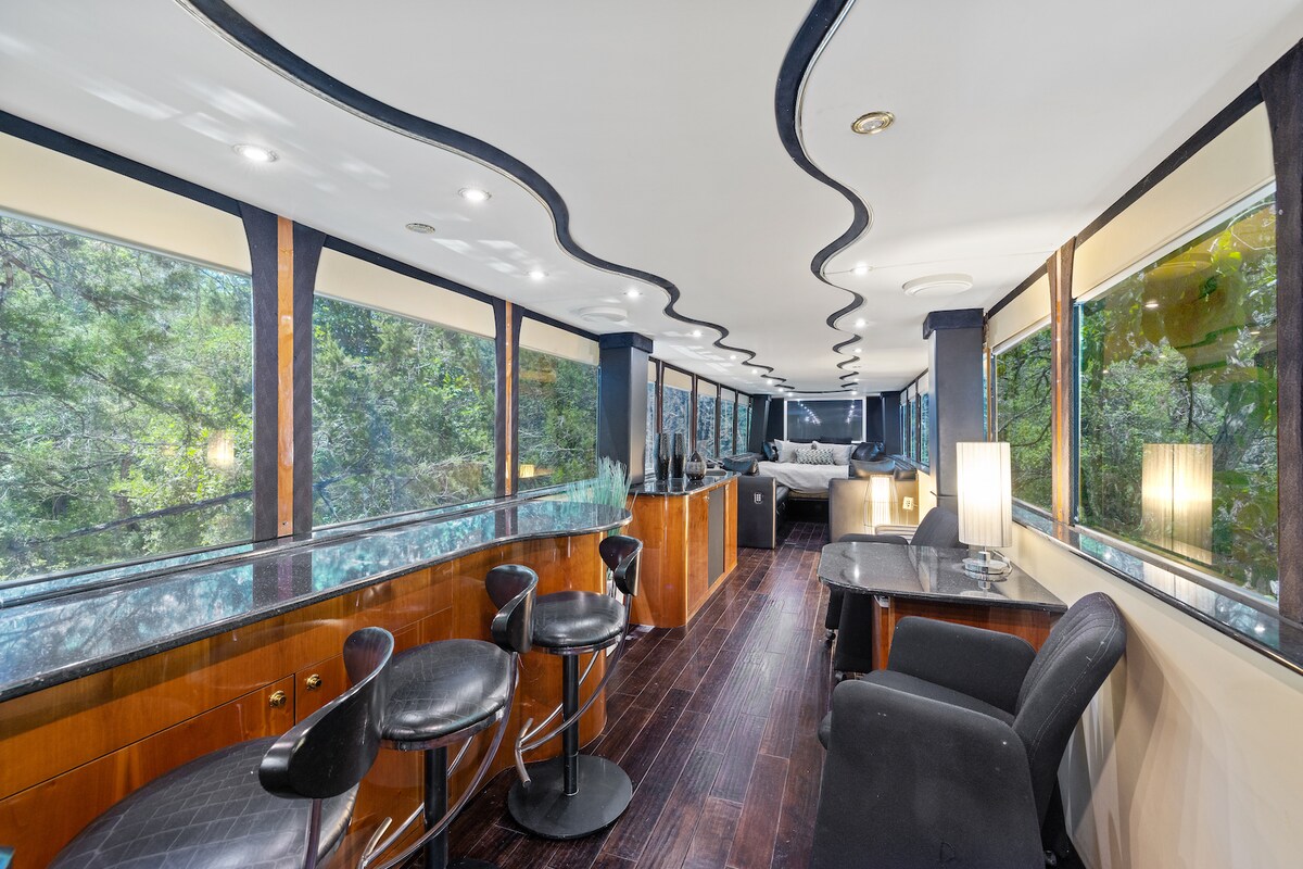 A spacious lounge area features sleek black seating, with a bar-style counter highlighted by granite surfaces. Expansive windows allow natural light to fill the space, offering views of the surrounding greenery. Modern furnishings create a comfortable atmosphere for relaxation or socializing.