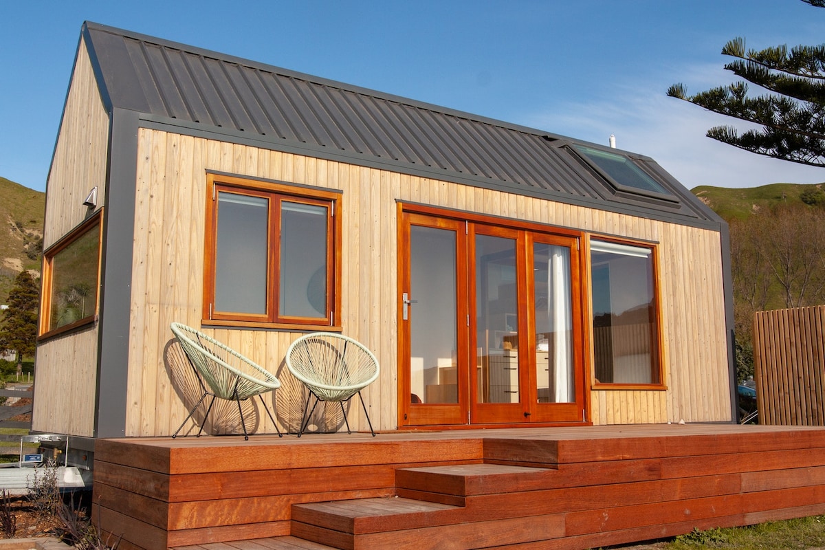The exterior of a modern wooden tiny house is showcased, featuring a sloped metal roof and large glass doors. Two stylish chairs are placed on the wooden deck, and a skylight above adds natural light. Surrounding hills can be seen in the background.