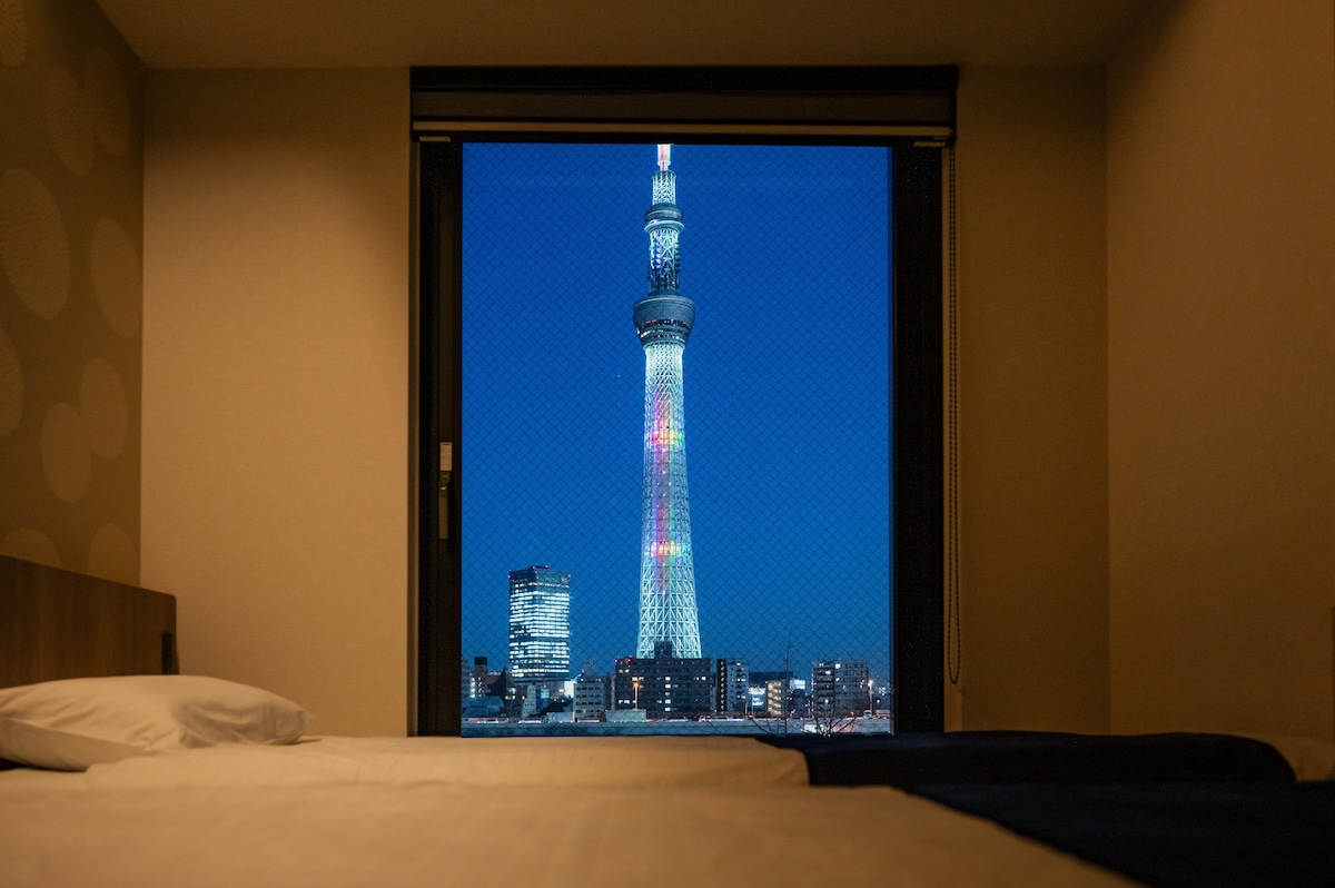 A large window offers a view of the Tokyo Skytree illuminated at night, showcasing vibrant colors against the dark sky. The spacious bed and light-colored linens create a calming setting, while city lights are visible in the background.