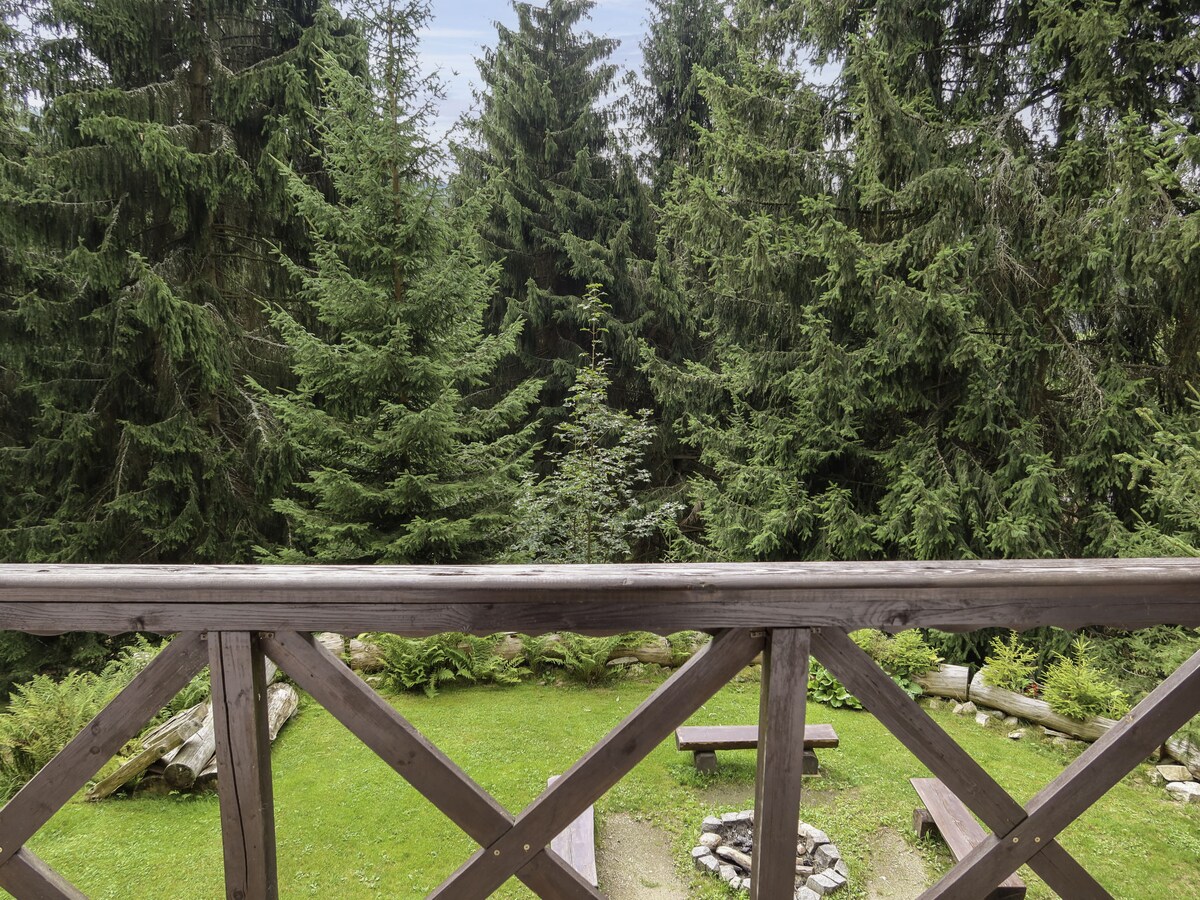 A balcony railing frames a lush greenery view, highlighting dense trees surrounding a grassy area. A fire pit is visible on the ground below, accompanied by wooden benches, creating a serene outdoor space for relaxation.
