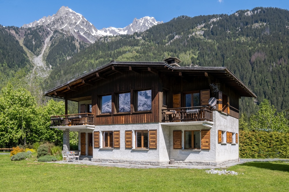 The chalet presents a two-story design, featuring large windows that showcase beautiful mountain views. A spacious lawn surrounds the property, complemented by a garden area. Balconies are visible on the upper level, providing outdoor seating options amidst the tranquil mountain landscape.