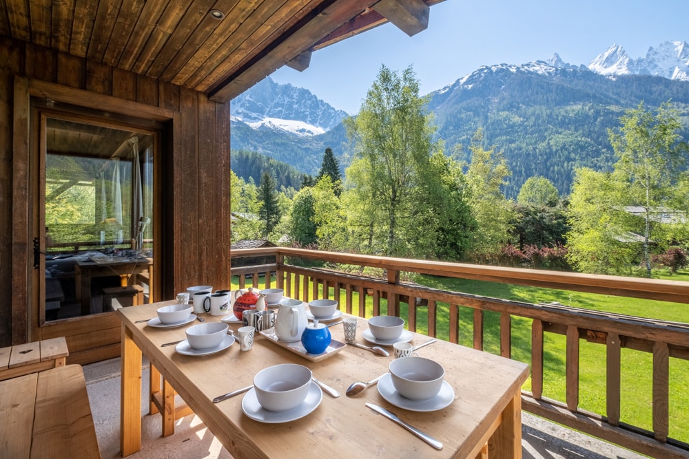 An outdoor terrace features a wooden table set for a meal, adorned with white plates and a kettle. Lush green gardens and majestic mountains provide a scenic backdrop, while large glass doors lead to the indoor dining area, inviting a seamless connection to nature.