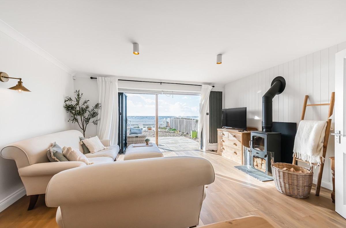 A light and airy living area features a comfortable seating arrangement, complemented by large windows that open to a beach view. A wood-burning stove provides warmth, while simple decor in neutral tones creates a contemporary feel. Access to the outdoor space is available through sliding doors.