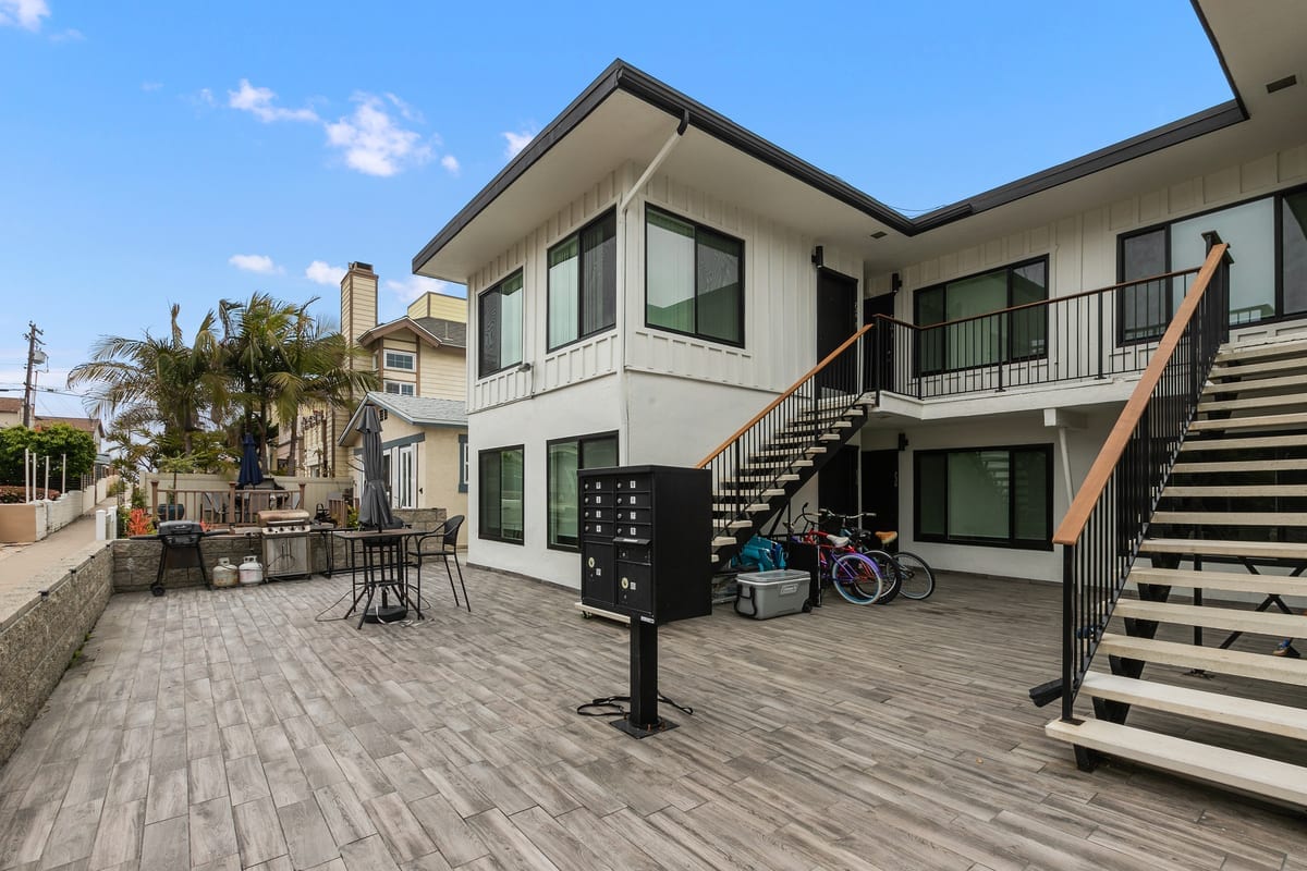 An outdoor area features a spacious patio with a wooden deck and modern seating arrangements. Sturdy stairs lead to the upper-level entrance, while a bike rack and storage containers are visible nearby. Lush greenery adds a touch of nature alongside the building.