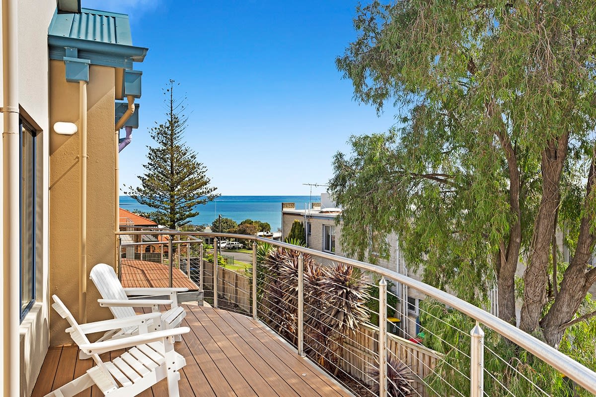 A spacious deck overlooks the ocean, offering views of the water and surrounding greenery. Two white lounge chairs are positioned on the wooden deck, inviting relaxation while enjoying the coastal scenery.