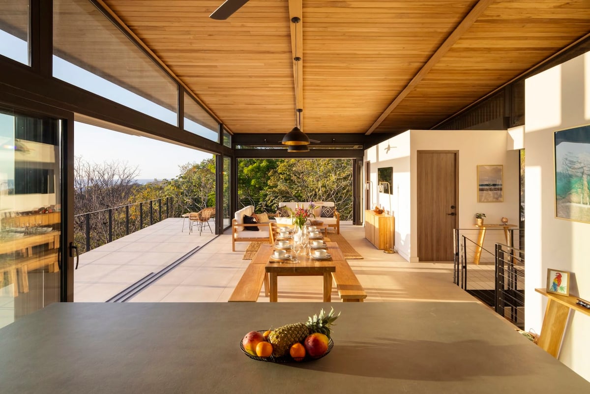 An open-concept dining area is featured, showcasing a long wooden table set for a meal. Large glass doors provide a seamless connection to the outdoor space, allowing natural light to illuminate the room. A countertop displays fresh fruits, adding a vibrant touch to the setting.