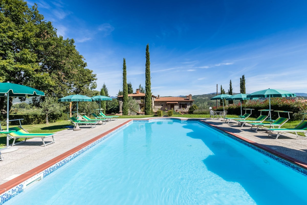 A large swimming pool is surrounded by sun loungers and parasols, set against a backdrop of manicured gardens and rolling hills. Towering cypress trees frame the scene, and the villa is visible in the distance under a clear blue sky.