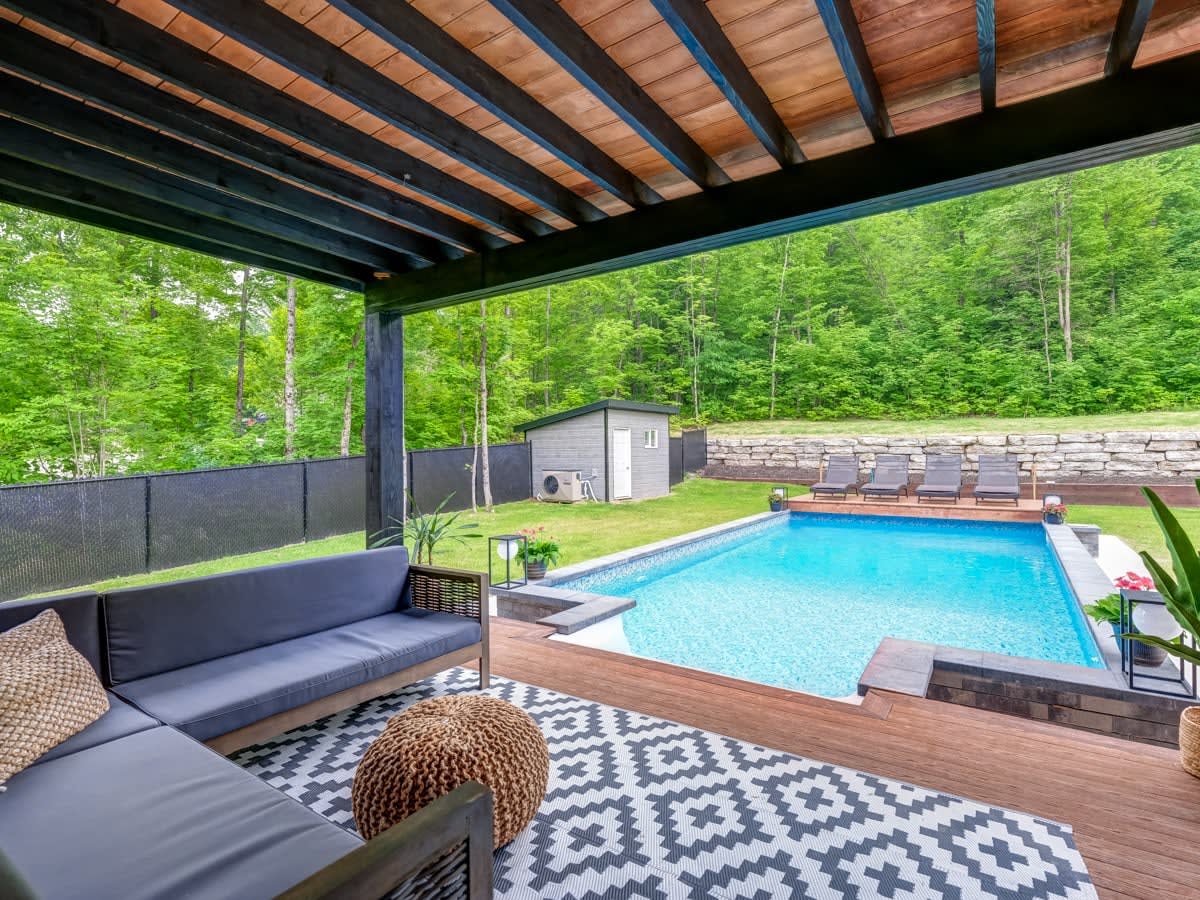 A covered outdoor seating area features a dark wooden ceiling and a cozy sofa set, complemented by a patterned rug. In the background, a bright blue pool is surrounded by loungers, with lush greenery creating a serene atmosphere.