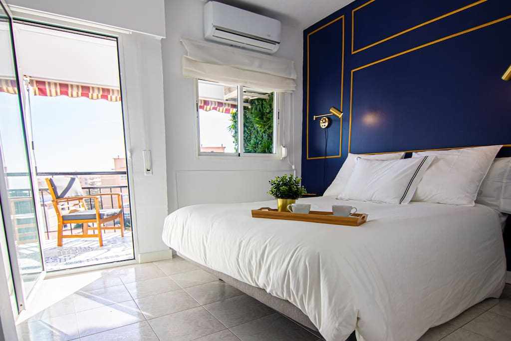 A modern bedroom features a spacious bed adorned with crisp white linens. A tray with cups is placed on the bed, and a large window opens to a private balcony. Soft natural light illuminates the room, complemented by a decorative blue wall.