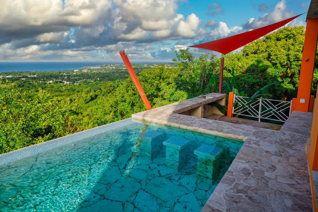An infinity pool is seen merging with the horizon, surrounded by natural stone and vibrant greenery. A slanted red awning provides shade over the poolside area, offering a spacious view of the landscape and distant ocean under a partly cloudy sky.