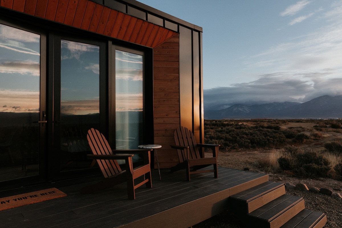 A charming deck features two wooden chairs and a small table, inviting relaxation while offering expansive views of the surrounding landscape. The sleek structure, with a blend of wood and modern materials, stands against a backdrop of mountains and a changing sky.