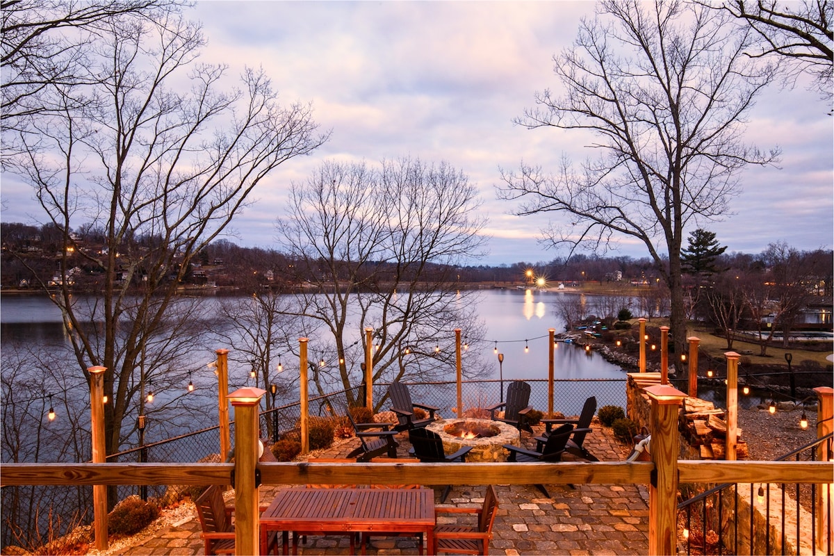 A stone patio is viewed overlooking a tranquil lake, featuring a fire-pit surrounded by comfortable seating. Soft evening light from string lights adds warmth, while trees frame the scenery, creating a serene atmosphere perfect for relaxing outdoors.