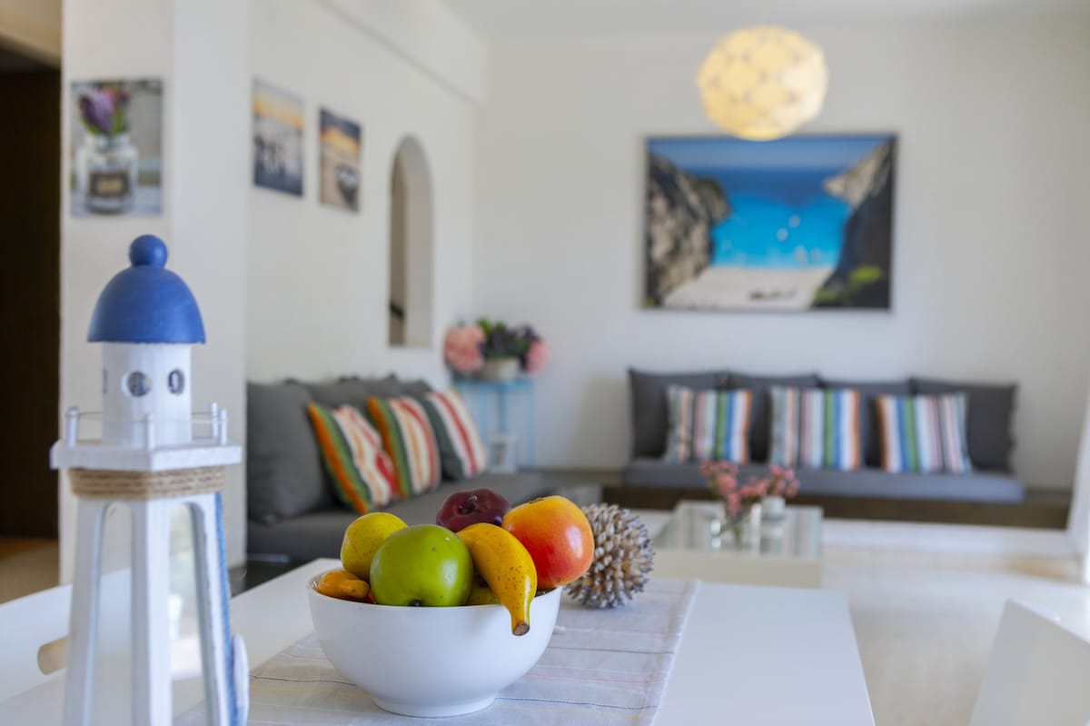 A dining table holds a bowl filled with various fruits, including apples, oranges, and bananas. In the background, a cozy seating area features gray sofas adorned with colorful striped cushions. A large artwork depicting a beach scene is prominently displayed on the wall, complemented by a decorative lighthouse figurine.