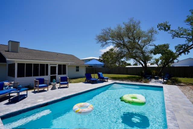 A fenced-in backyard features a large, heated swimming pool adorned with colorful flotation devices. Several lounge chairs are positioned around the pool, and shaded seating is provided under a blue umbrella. Lush greenery complements the serene outdoor space.