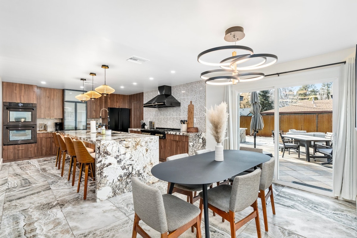 The modern kitchen features a large island with seating for four and a dining table surrounded by six chairs. High-end appliances are highlighted, including dual ovens and a stylish hood. Natural light fills the space through sliding glass doors that open to the outdoor patio.