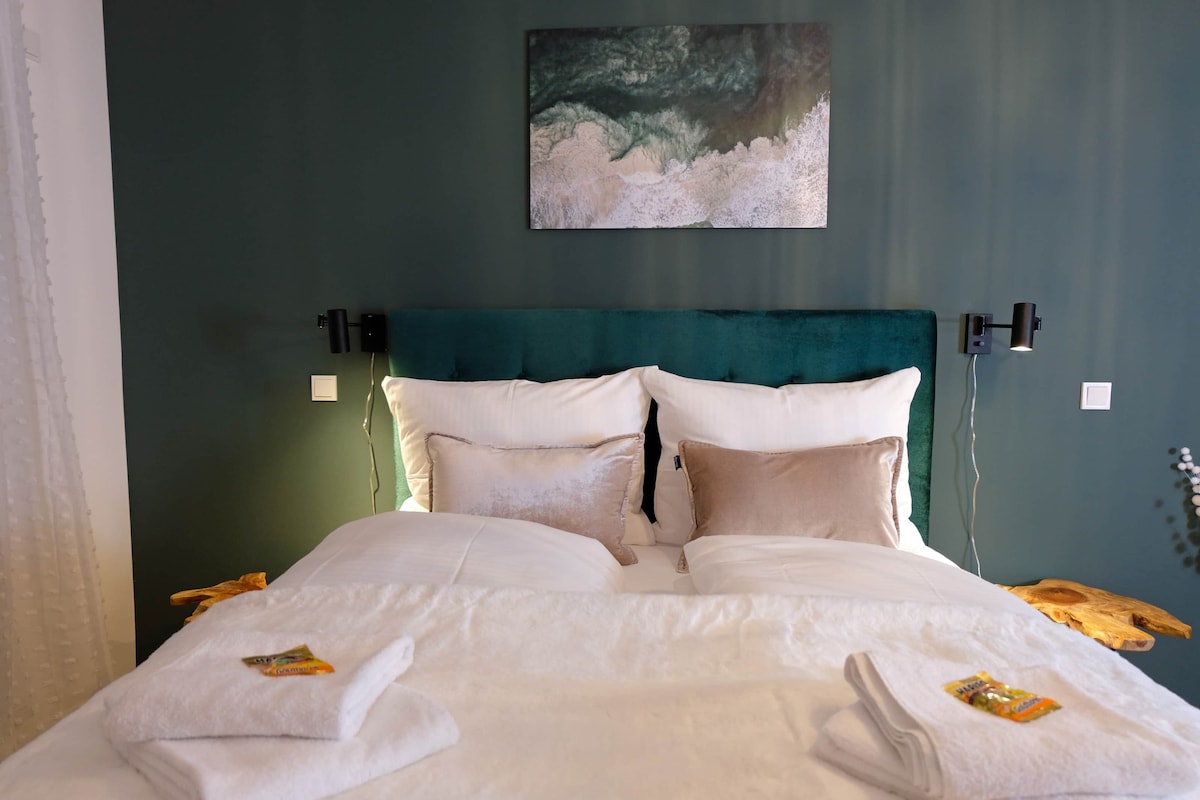 A comfortable sleeping area features a deluxe king-size bed adorned with plush pillows and soft bedding. A textured green accent wall serves as a backdrop, while two minimalist light fixtures are mounted at either side. Fresh towels are neatly arranged at the foot of the bed.
