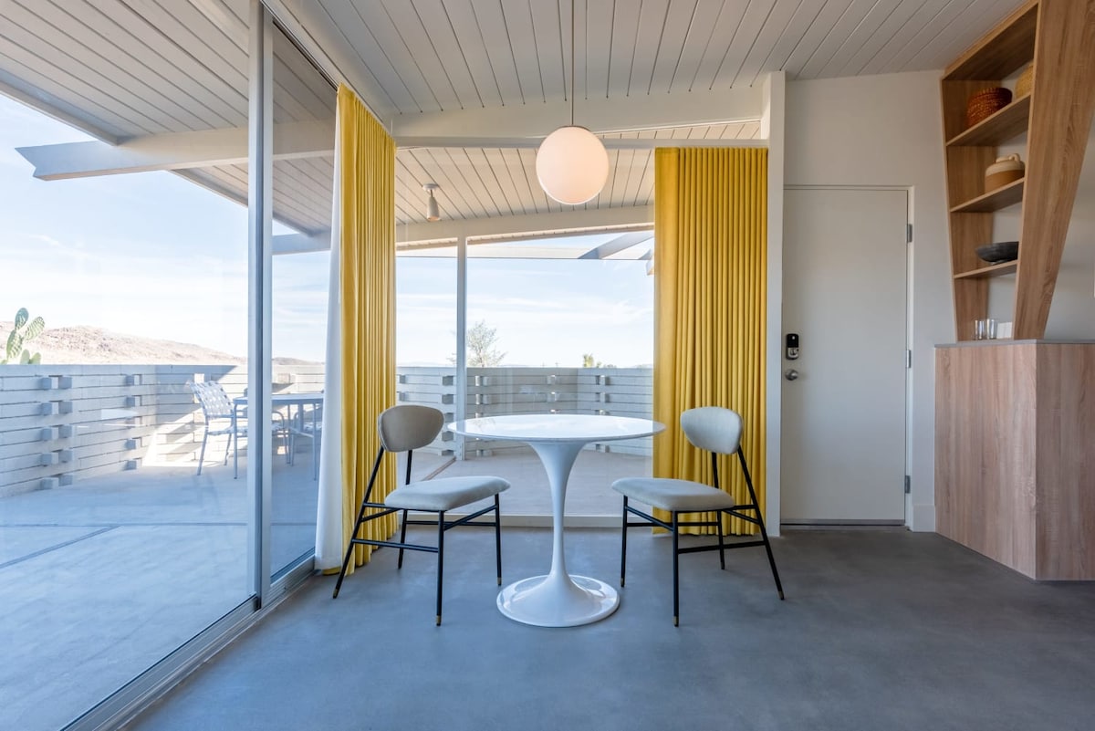 A sunlit dining area is defined by a round white table surrounded by two light grey chairs. Floor-to-ceiling glass panels offer views of the outdoors, framed by vibrant yellow curtains. A modern light fixture hangs above, complementing the sleek design.