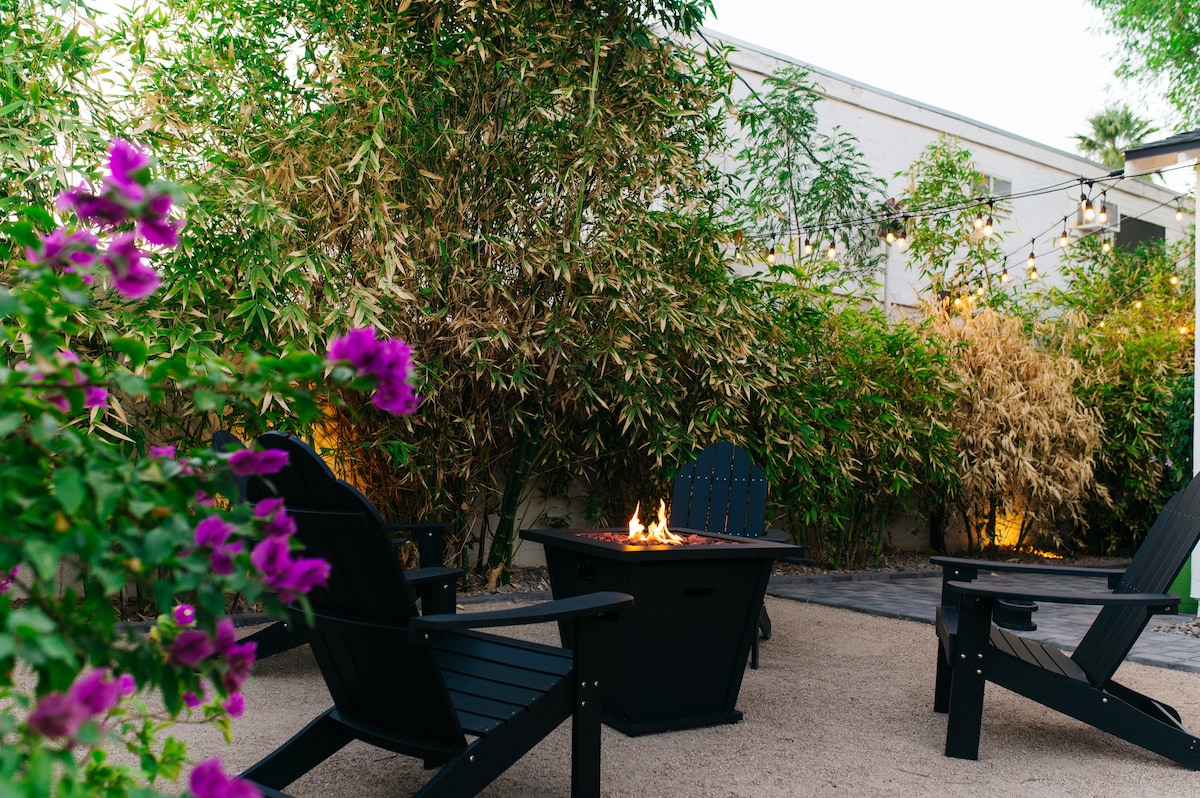 A cozy outdoor space features two black Adirondack chairs positioned around a fire pit, which has a gentle flame. Lush greenery and flowering plants provide a serene backdrop, enhancing the inviting atmosphere of the enclosed yard.