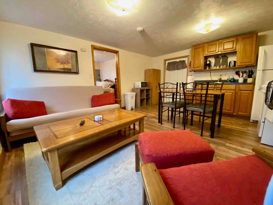 A welcoming living area features a futon and two red accent chairs facing a wooden coffee table. In the background, a dining table with chairs is situated adjacent to a fully stocked kitchen, showcasing wood cabinetry. Soft lighting enhances the warm tone of the space.