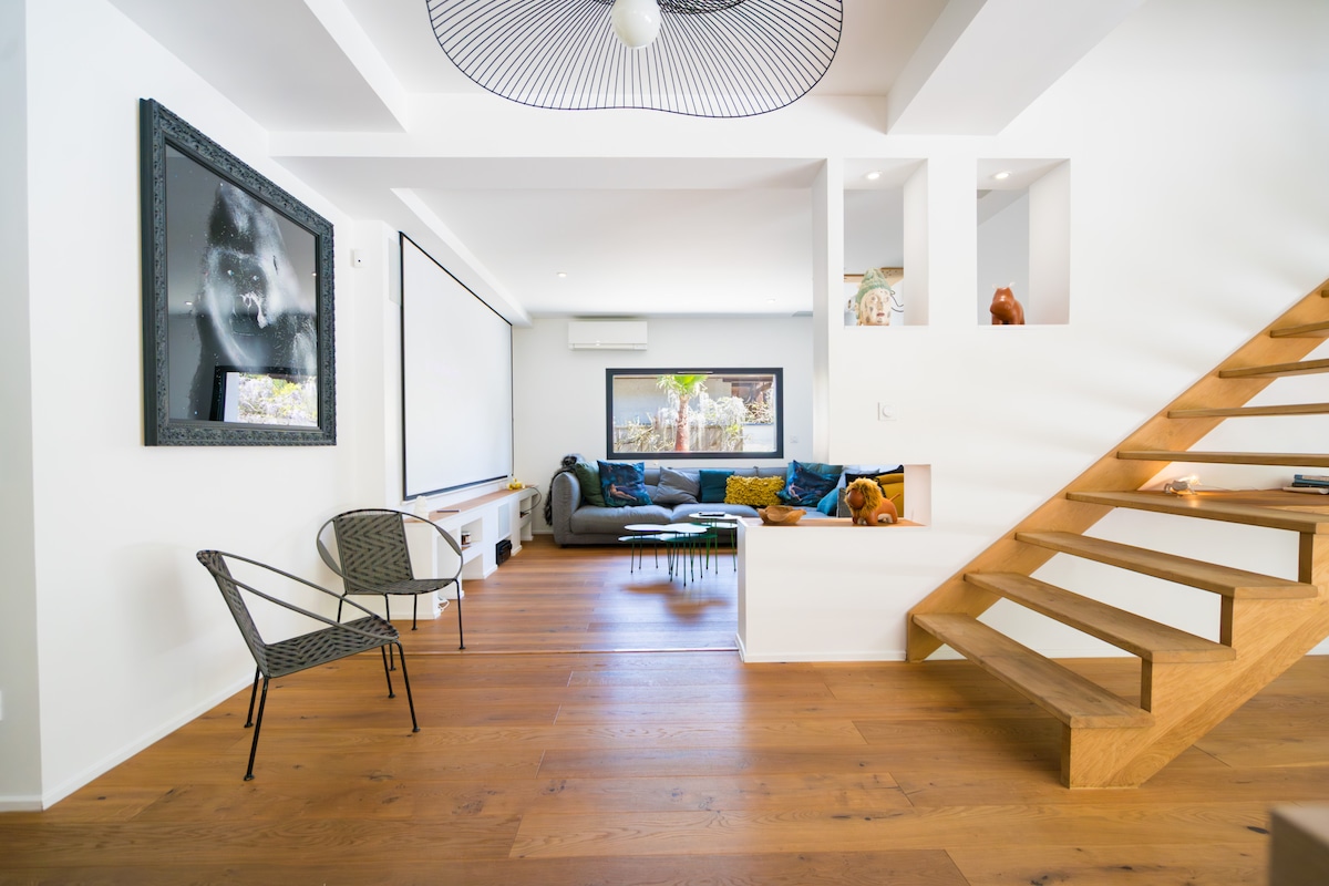 A bright and spacious living area features wooden flooring, a comfortable grey sofa adorned with colorful cushions, and a large screen positioned on the wall. An elegant staircase leads to the upper level, and a modern light fixture hangs above the area.
