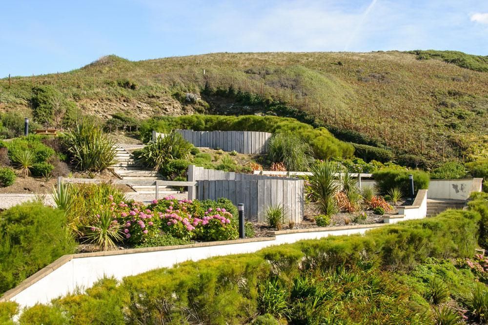 Manicured communal gardens feature a pathway leading through a variety of lush greenery and blooming flower beds. Elevated landscaping creates tiered terraces with wooden fencing. The gentle slope in the background complements the serene environment.