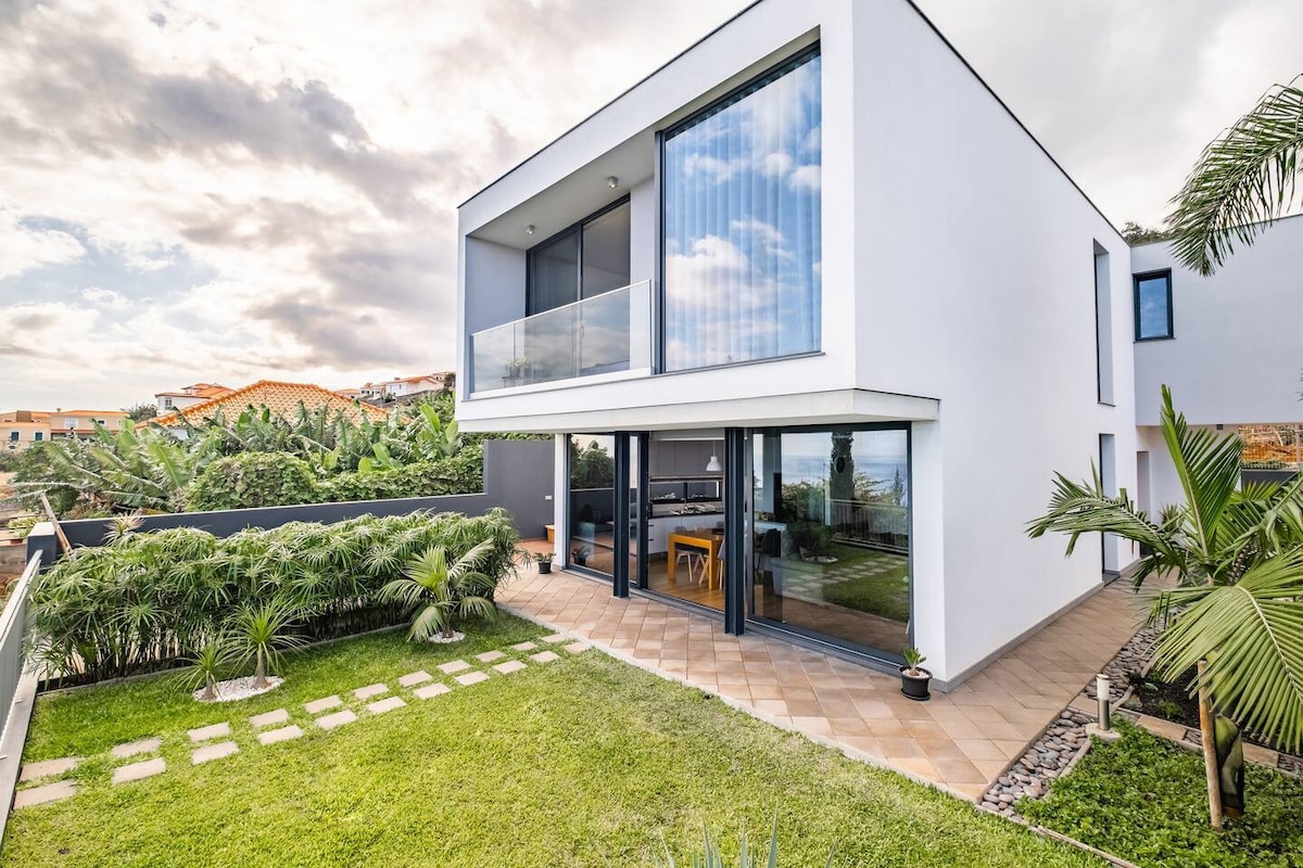 A modern two-story house features large glass windows and a spacious outdoor area. The lush garden is surrounded by greenery, with a stone path leading to the entrance. The design seamlessly blends indoor and outdoor spaces, showcasing an inviting atmosphere.