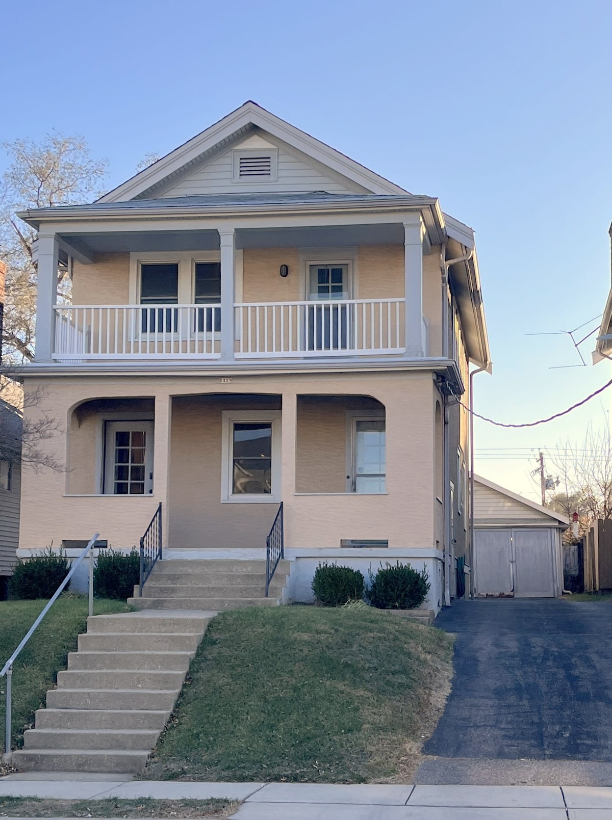 A well-maintained two-story house is viewed from the front, featuring a beige exterior and a covered porch with white railings. Steps lead up to the entrance, with a grassy area and a paved driveway leading to a garage at the side.