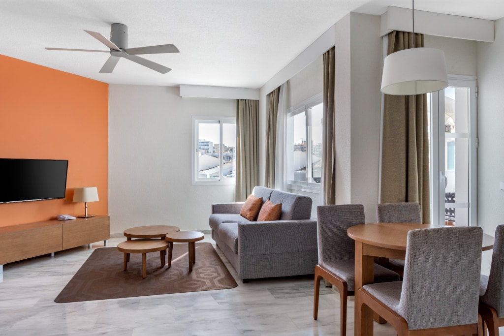 A bright living area is highlighted by an orange accent wall and light-colored flooring. It features a comfortable sofa, two coffee tables, and a dining table with chairs. Large windows allow for natural light, complemented by soft curtains. A ceiling fan adds a modern touch.