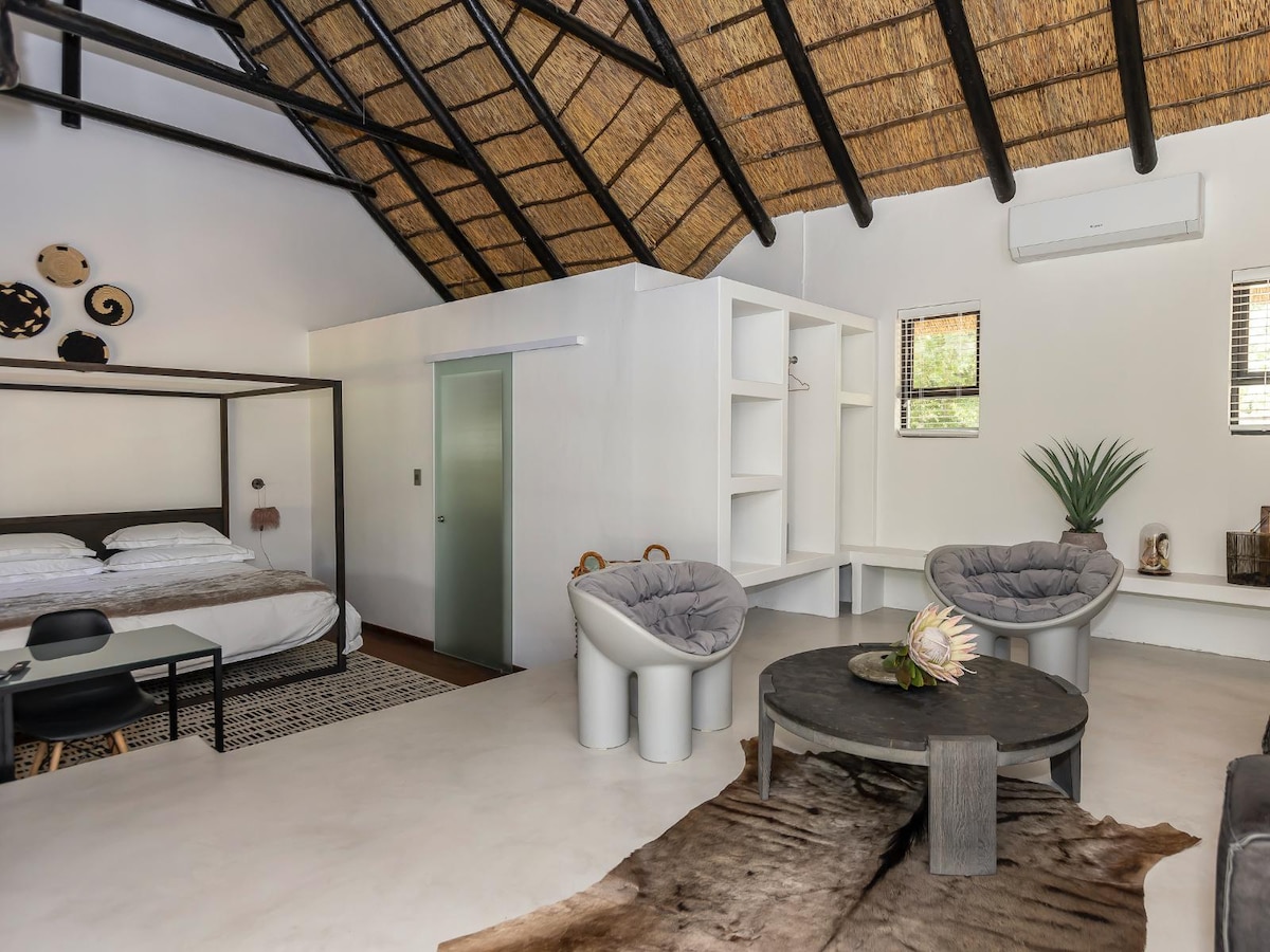 The spacious studio features a 4-poster king-size bed, complemented by wooden flooring. A circular table is surrounded by unique chairs, creating a cozy sitting area. Natural light enters through windows, and a decorative plant adds a touch of greenery to the modern decor.
