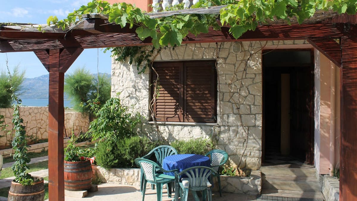 A charming outdoor seating area is featured under a wooden pergola, offering shade and views. A blue table is surrounded by green chairs. Vines and foliage adorn the space, and stone walls complement the natural surroundings. The entrance to the apartment is visible to the right.