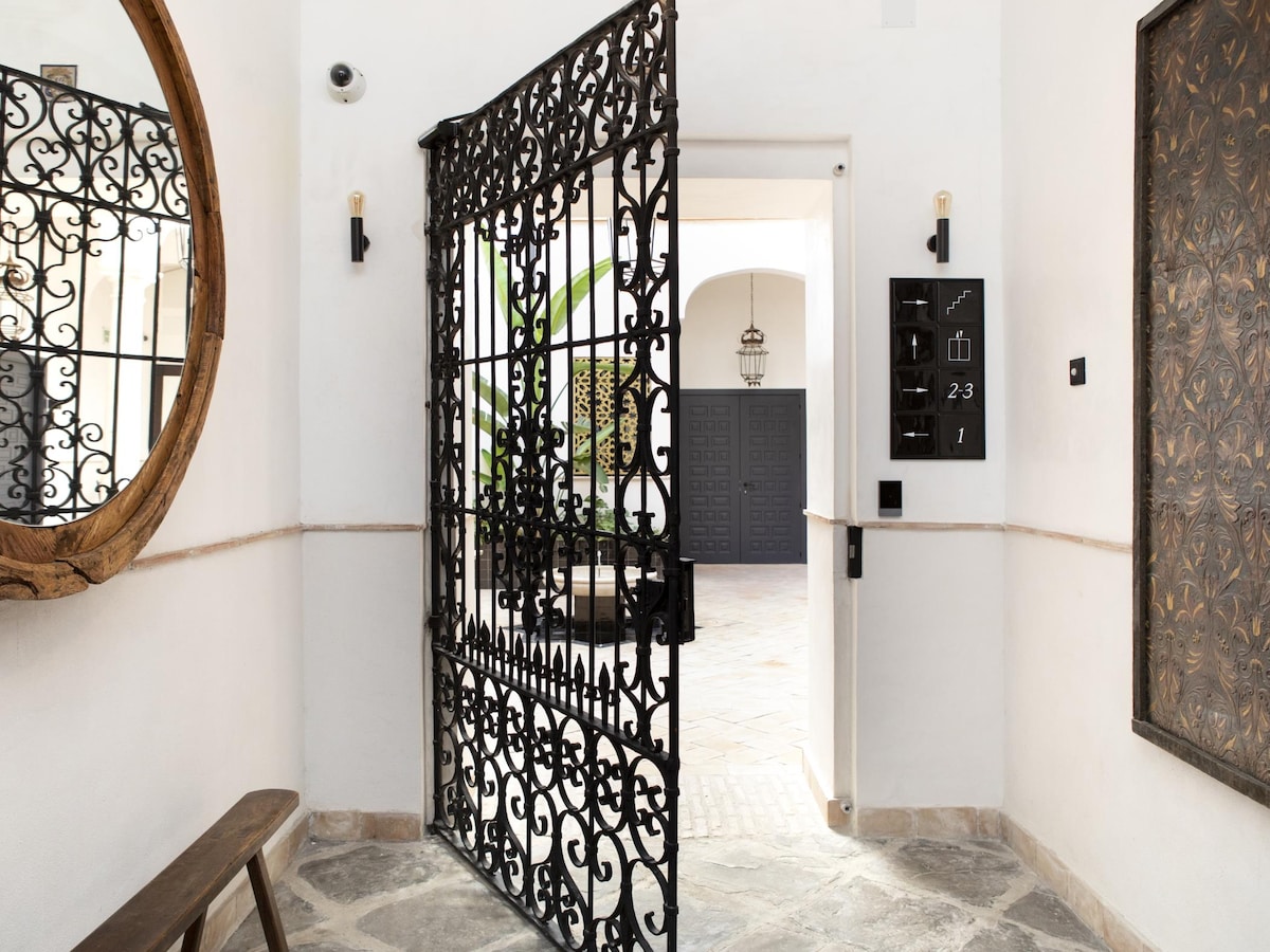 An intricate iron gate is showcased, opening to a bright interior patio featuring a stone floor. A round mirror reflects the entrance area, where a wooden bench is placed against the wall. The space is enhanced by overhead lighting and greenery visible in the courtyard.