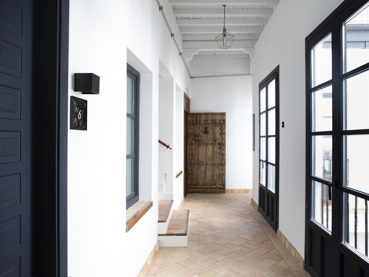 A corridor features a serene layout with white walls and terracotta tiled flooring. Wooden doors and windows provide natural light, while a black number six plaque indicates the apartment's location. The ambiance is complemented by a rustic wooden door at the end of the hallway.