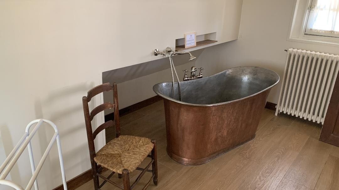 A vintage-style bathroom features a stylish, oval copper bathtub positioned against a wall. Beside it, a wooden chair with a woven seat is placed. A sleek radiator is visible on the adjacent wall, contributing to the room's overall warmth.
