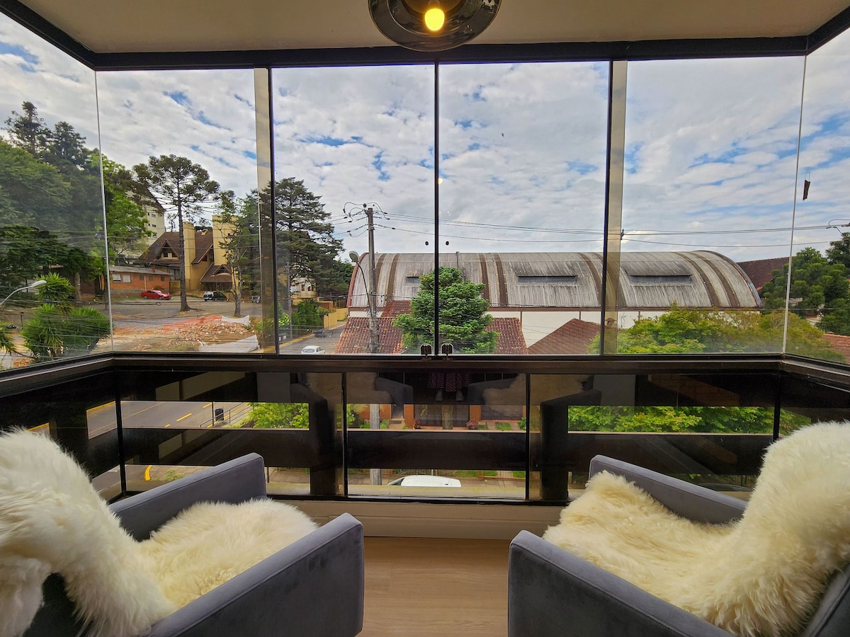 A bright balcony area features large glass panels offering an unobstructed view of the surrounding neighborhood. Two comfortable gray chairs with white throw blankets are positioned to invite relaxation while enjoying the scenery outside.