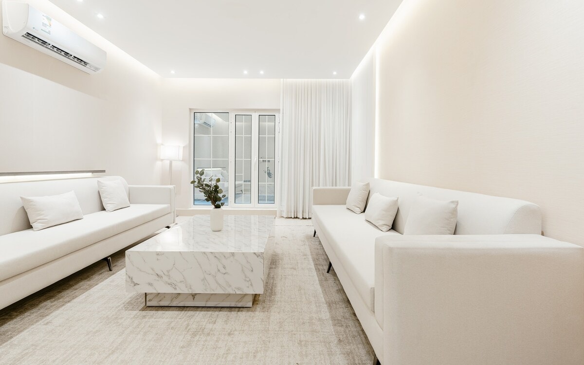 A modern living room is featured, showcasing two sleek white sofas arranged around a marble coffee table. Natural light filters through large glass doors, creating a bright atmosphere. Soft curtains frame the window, while minimalistic decor enhances the sense of space.