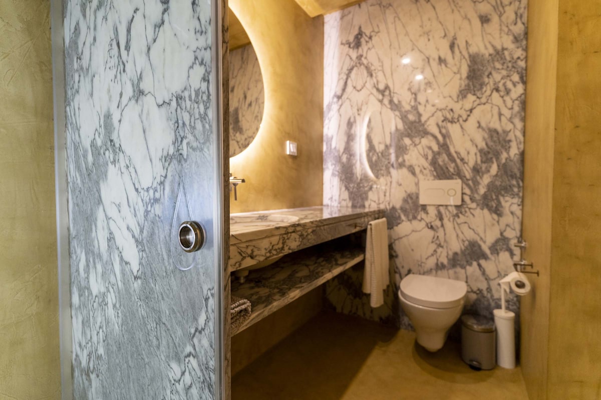 A marble bathroom features a round mirror above a countertop with an integrated sink. The walls display a marbled pattern in grey and white. A toilet is positioned nearby, with a roll of toilet paper and a wastebasket easily accessible.