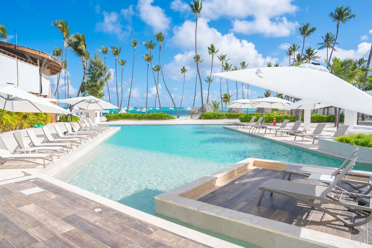 A large swimming pool is surrounded by sun loungers under white umbrellas, with palm trees swaying gently in the breeze. The clear blue water of the pool reflects the sunny sky and hints at the beachfront in the background.