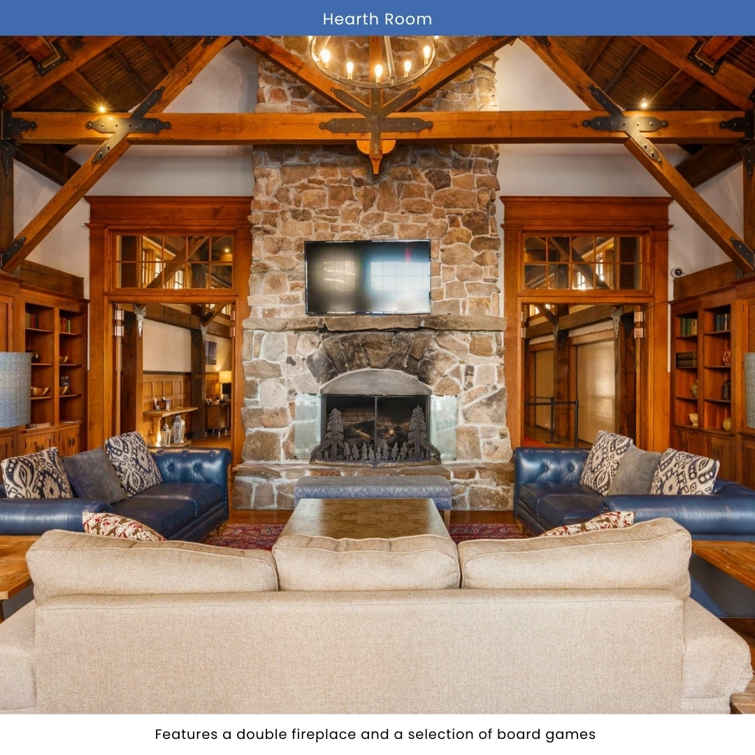 The Hearth Room displays a double fireplace framed by a stone wall, providing a cozy focal point. Seating includes a large, light-colored sofa and dark leather chairs. A selection of board games is available on a central wooden table, inviting guests to gather together.