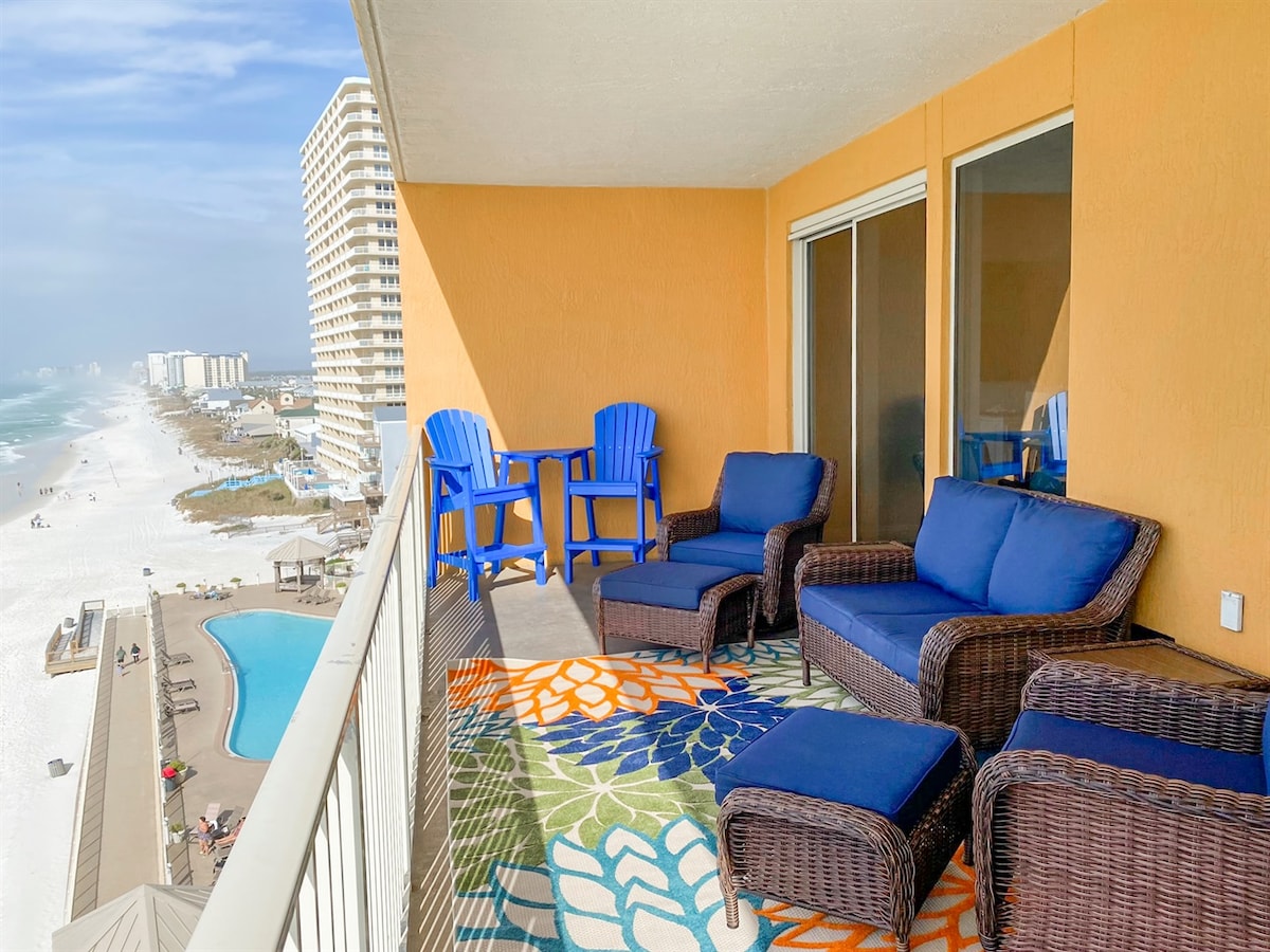 A spacious balcony is furnished with comfortable seating, including two chairs and a loveseat, all set against a vibrant outdoor rug. A table complements the arrangement, offering a perfect spot to enjoy ocean views and fresh air.