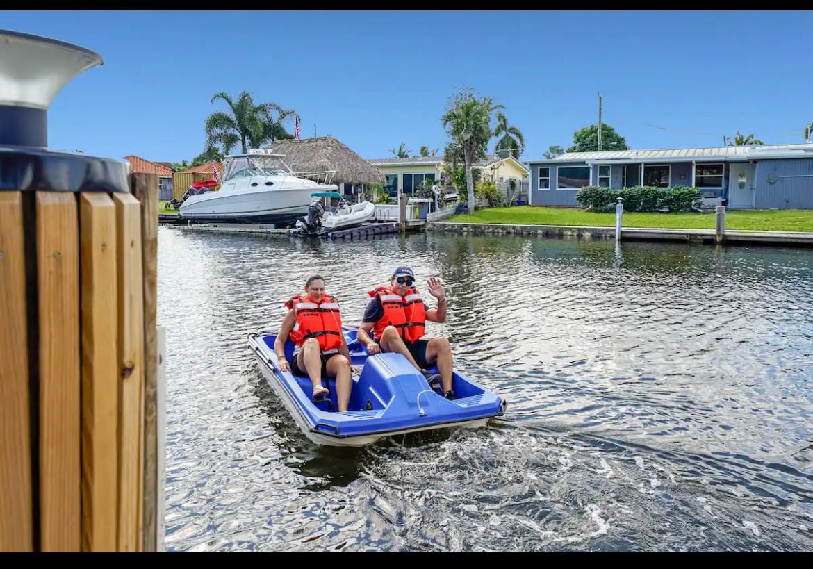 A pedal boat is seen gliding along the calm waters of a canal, with two individuals wearing life jackets enjoying their ride. Nearby, various boats are docked along the waterfront properties, surrounded by palm trees and well-maintained yards.