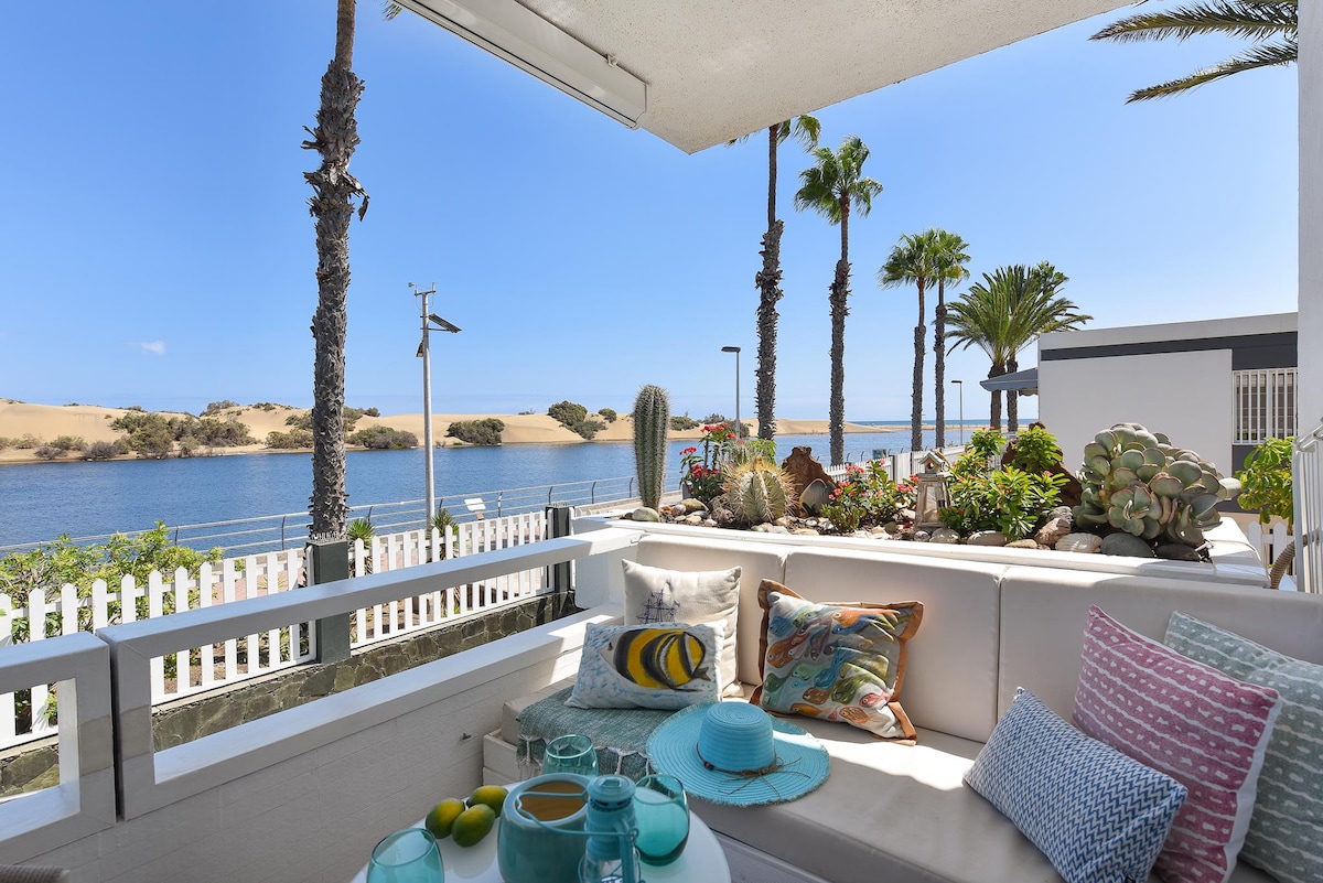 A comfortable outdoor seating area is positioned on a terrace, featuring plush cushions and a coffee table. A view of palms and a scenic lagoon is visible in the background, bordered by sandy dunes under a clear blue sky. Decorative plants enhance the relaxing environment.